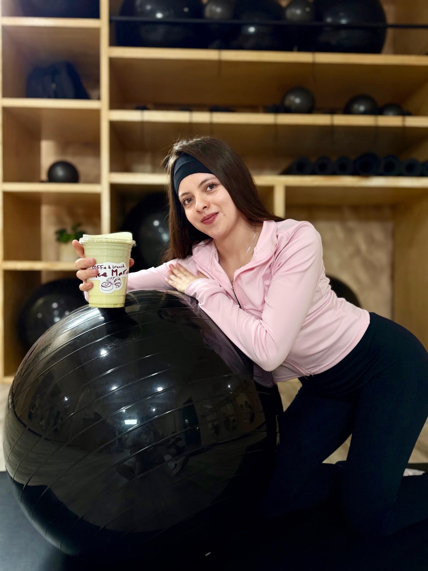 Alumna sonriente de YW Studio recargada en una pelota de pilates con su bebida en mano