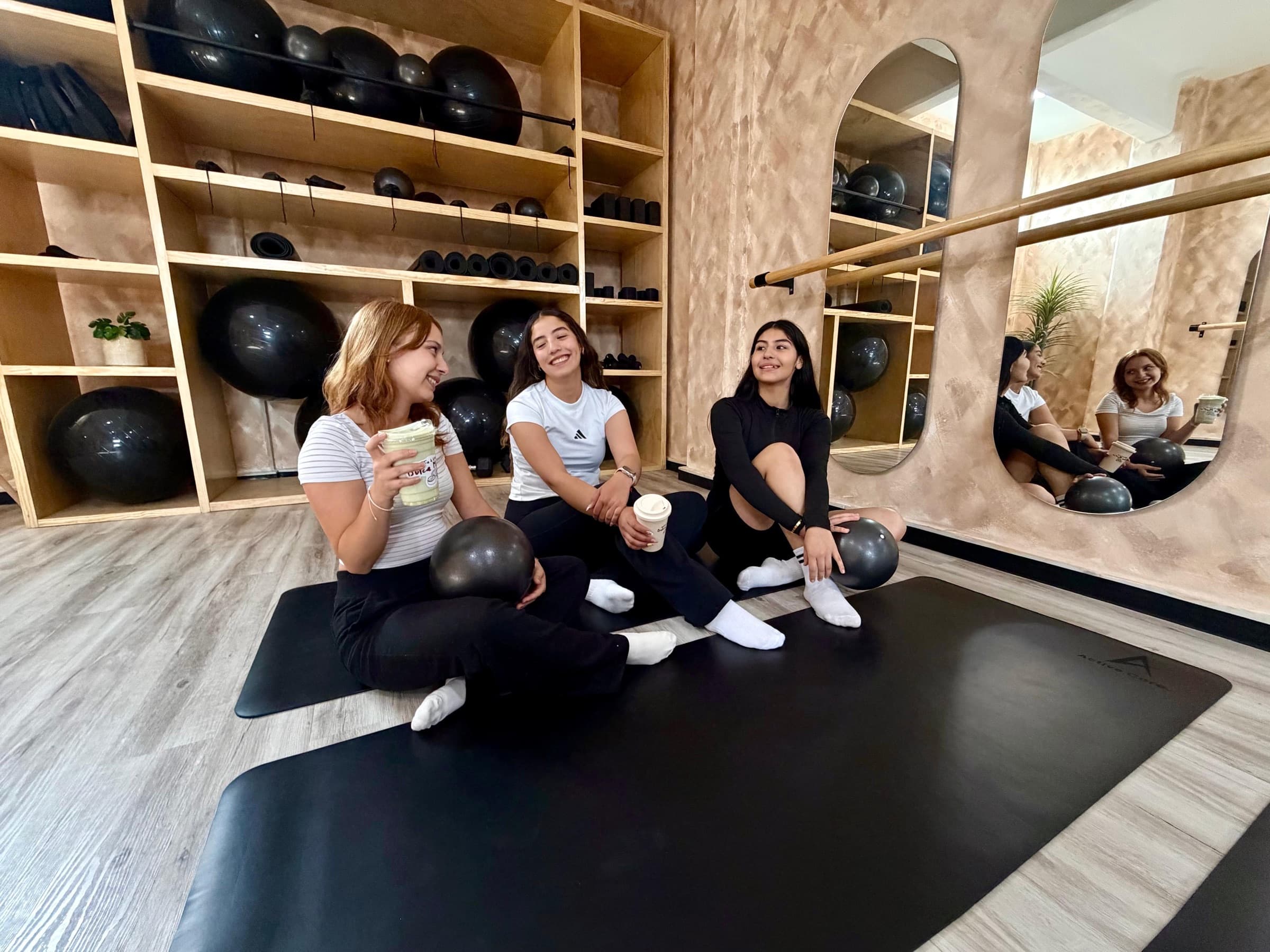 Alumnas de YW Studio conversando sobre tapetes en el salón con repisas de pelotas de pilates y espejos en arco